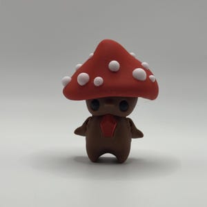 May include: A charming mushroom figure with a red cap and white spots. The figure has a brown body, small arms, and a red gem-like shape on its chest. The eyes are black and round.