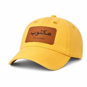 Könnte beinhalten: Senfgelbe Baseballkappe mit gebogenem Schirm. Ein brauner Lederaufnäher auf der Vorderseite zeigt arabischen Text und den englischen Satz "it's written". Die Kappe hat ein klassisches Sechs-Panel-Design.