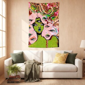 May include: A colorful tapestry featuring a surreal illustration of a woman with multiple eyes and faces. The artwork uses shades of green, pink, and red, with paisley and floral patterns in the background. The tapestry is hanging on a beige wall above a white sofa.