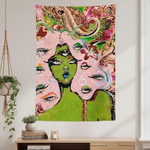May include: A tapestry featuring a vibrant, surreal illustration of a woman with multiple eyes and faces. The artwork uses a palette of pink, green, and brown, with paisley and floral patterns in the background. The central figure has green skin and blue lips.