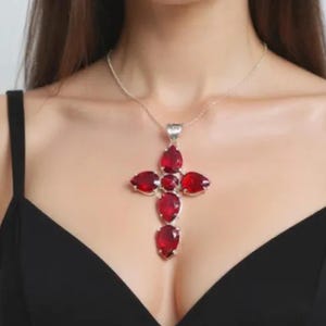 Pode incluir: Um colar prateado com um grande pendente de cruz. A cruz é adornada com múltiplas pedras preciosas vermelhas escuras em forma de gota. O colar é usado sobre um top preto.