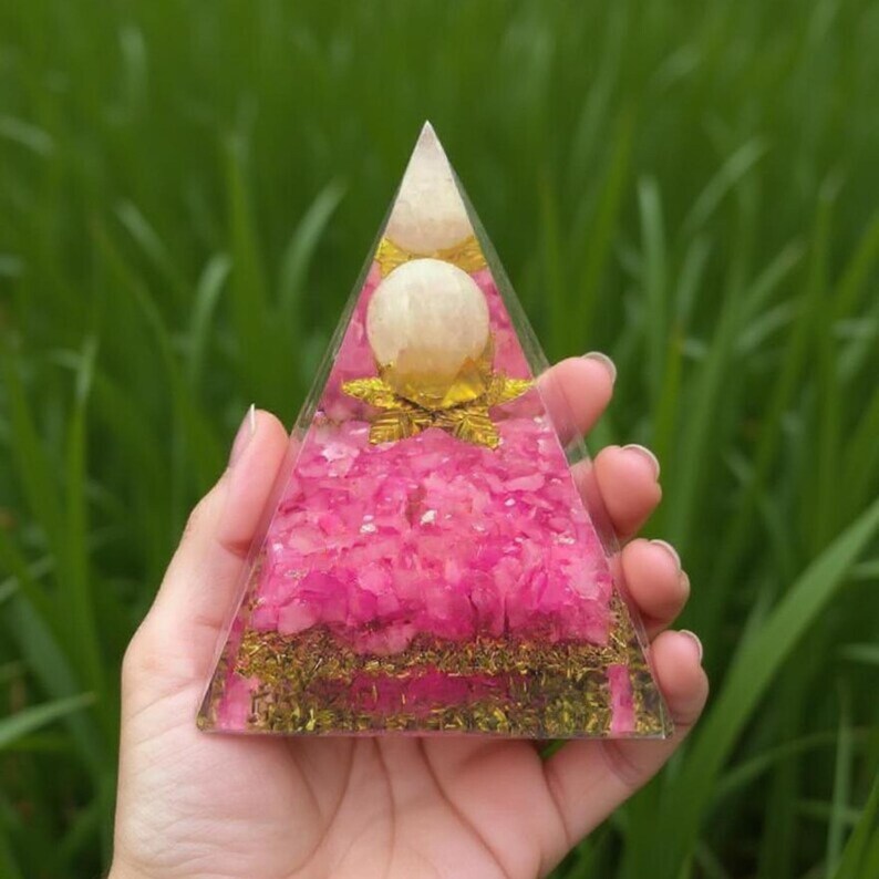 A Rose Quartz pyramid held in a person's hand to show the 3-inch scale and 200-300g weight. The smooth, polished surface reflects the light, making it a perfect tactile object for meditation, prayer, or focused breathing exercises.