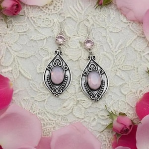 May include: A pair of silver-toned dangle earrings featuring a teardrop-shaped frame with intricate scrollwork detailing. Each earring is centered with an oval-cut pale pink stone and topped with a smaller round-cut pink stone on the hook.