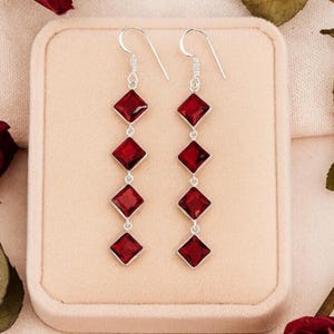 Puede incluir: Un par de pendientes largos con cuatro piedras de color rojo intenso, talladas en forma cuadrada y engastadas en metal plateado. Cada pendiente cuelga de un gancho plateado, con las piedras dispuestas verticalmente en un patrón lineal.