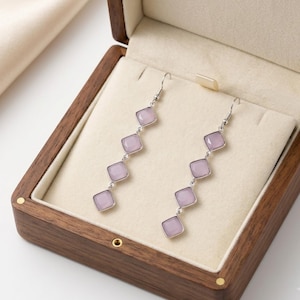 May include: A pair of silver-toned drop earrings featuring four square-shaped, light purple stones arranged in a vertical line. Each stone is set in a metallic frame, connected by small silver links, and finished with a standard hook-style earring wire.