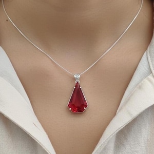 Op de afbeelding: Een zilveren ketting met een grote, facetgeslepen, rode hanger. De hanger heeft een dieprode kleur en een geometrische vorm. De ketting wordt gedragen op een wit overhemd.