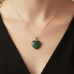 May include: A silver necklace featuring a heart-shaped pendant. The pendant displays a blend of green and purple colours. The necklace is set against a black top.