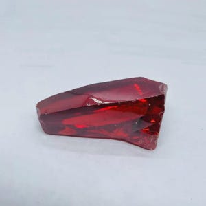 Könnte beinhalten: Ein facettierter, tiefroter Glaskristall mit unregelmäßiger Form. Der Kristall ist durchscheinend, lässt Licht durch und hat eine leicht glänzende Oberfläche. Die Farbe ist ein leuchtendes, sattes Rot.