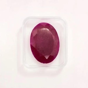 May include: A faceted, oval-shaped ruby gemstone in a clear plastic case. The ruby is a deep, rich red color, with visible internal facets. The case is square with rounded corners, set against a white background.