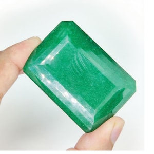 Pode incluir: Uma grande pedra preciosa retangular verde esmeralda com bordas facetadas. A gema é segurada entre os dedos, mostrando sua superfície polida e sua cor vibrante. A forma da gema é um retângulo com bordas chanfradas.