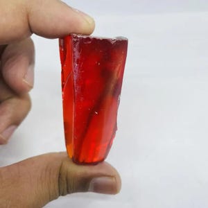 Può includere: Una gemma rettangolare traslucida rosso-arancio è tenuta tra le dita. La gemma ha una superficie ruvida e irregolare e un aspetto lucido. Il colore passa da un rosso intenso a una tonalità arancione più chiara, con strutture interne visibili.