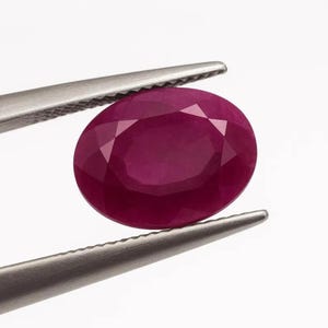 May include: A faceted, oval-shaped ruby gemstone held by silver tweezers. The ruby is a deep, rich red color, with visible facets that catch the light. The tweezers are made of a shiny, metallic material.