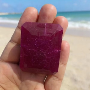 May include: A large, faceted, rectangular ruby gemstone with a deep magenta color. The gem is held in a hand, with a beach and ocean in the background. The ruby's surface displays internal veining.