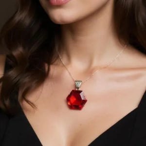 May include: A gold-coloured necklace featuring a large, faceted, red gemstone pendant. The pendant is hexagonal in shape. The necklace is worn on a person.