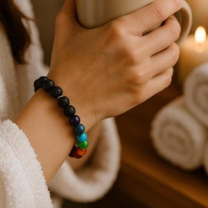 May include: A bracelet made of black lava stones and seven chakra beads in rainbow colours. The bracelet is worn on a wrist. The beads are red, orange, yellow, green, blue, indigo, and purple. The bracelet is a mix of dark and vibrant colours.