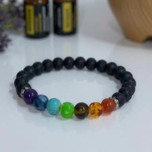 Op de afbeelding: Een zwarte lavasteen armband met zeven chakra kralen in verschillende kleuren. De kralen zijn onder andere paars, blauw, turquoise, groen, bruin, oranje en rood. De armband ligt op een witte ondergrond.