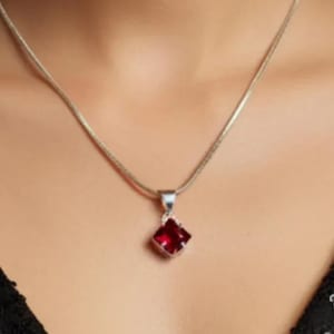 Peut inclure: Un collier en argent avec un pendentif carré rouge foncé. Le pendentif est serti dans un cadre argenté et est suspendu à une fine chaîne argentée. Le collier est présenté sur un fond de teint.