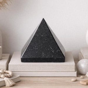 May include: A black granite pyramid with a speckled texture, resting on a stack of neutral-toned books. The pyramid's geometric shape and dark color create a striking contrast. Decorative item for home decor.