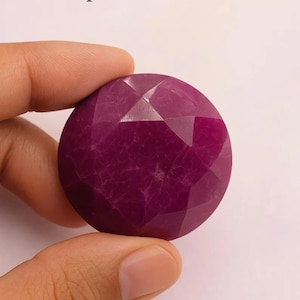 Peut inclure: Une pierre précieuse de forme ronde, taillée en facettes, de couleur rouge foncé. La gemme présente des inclusions internes visibles. La pierre est tenue entre les doigts, mettant en valeur sa taille et sa forme. La taille crée de multiples surfaces réfléchissantes.