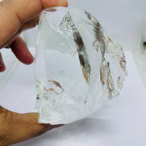 May include: A large, clear quartz crystal with a rough, irregular shape. The crystal is held in a hand, showcasing its translucent quality and natural texture. The crystal appears to be a raw, uncut specimen.