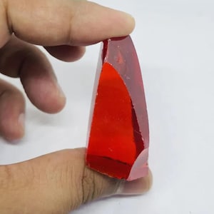 May include: A faceted, translucent, red gemstone is held between fingers. The gem has a triangular shape with a flat base and a pointed top. The edges are slightly rough, and the gem appears to be polished.
