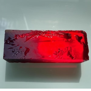 Peut inclure: Une pierre précieuse rectangulaire et translucide d'un rouge profond. La pierre a une surface rugueuse et texturée avec des zones de coloration plus foncée, créant un effet de dégradé. Il s'agit probablement d'un rubis ou d'une pierre précieuse similaire.