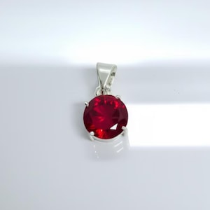 May include: A silver pendant featuring a large, round, deep red gemstone. The gemstone is held in place by four prongs, and the pendant has a simple, elegant design. The pendant is set against a white background.