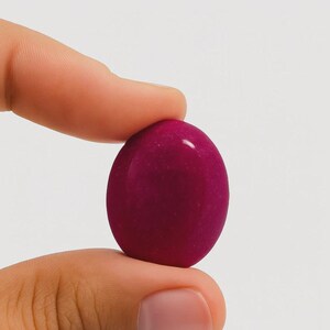 Peut inclure: Une pierre lisse, ovale, de couleur magenta foncé est tenue entre deux doigts. La pierre a une surface brillante et réfléchit la lumière. Le fond est de couleur blanc cassé.