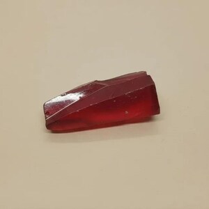 Pode incluir: Um objeto retangular facetado e translúcido em um tom vermelho profundo. O objeto tem uma forma geométrica com múltiplos planos e ângulos, que capturam a luz. A aparência geral sugere uma peça decorativa ou artística.