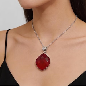 Op de afbeelding: Een zilverkleurige ketting met een grote, facetgeslepen, rode edelsteen hanger. De hanger heeft een diamantvorm en de ketting is een delicate schakelstijl. De ketting wordt gedragen over een zwarte top.