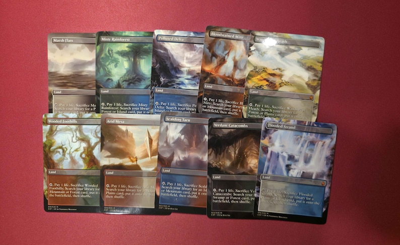 Pode incluir: Uma cole&ccedil;&atilde;o de cartas colecion&aacute;veis Magic: The Gathering, apresentando v&aacute;rias cartas de terreno com arte de paisagens. As cartas exibem nomes como "Marsh Flats" e "Verdant Catacombs" com ilustra&ccedil;&otilde;es e texto detalhados. As cartas est&atilde;o dispostas em uma superf&iacute;cie vermelha.