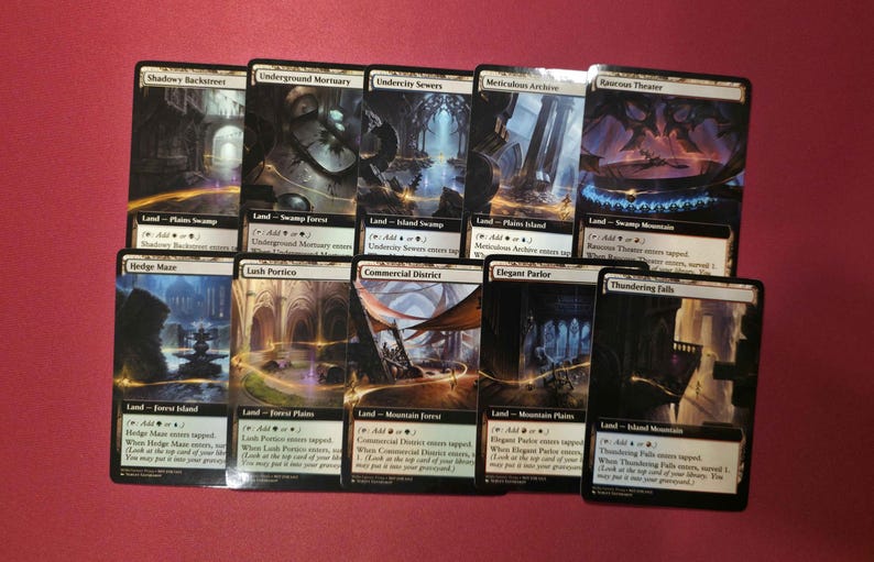 Pode incluir: Uma cole&ccedil;&atilde;o de dez cartas colecion&aacute;veis Magic: The Gathering. Cada carta apresenta obras de arte de fantasia e texto &uacute;nicos, com t&iacute;tulos como "Shadowy Backstreet" e "Underground Mortuary". As cartas est&atilde;o dispostas sobre uma superf&iacute;cie vermelha.