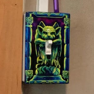 May include: A decorative light switch cover featuring a green gargoyle design against a purple and black background. The cover has a blue border with green accents and a white switch.