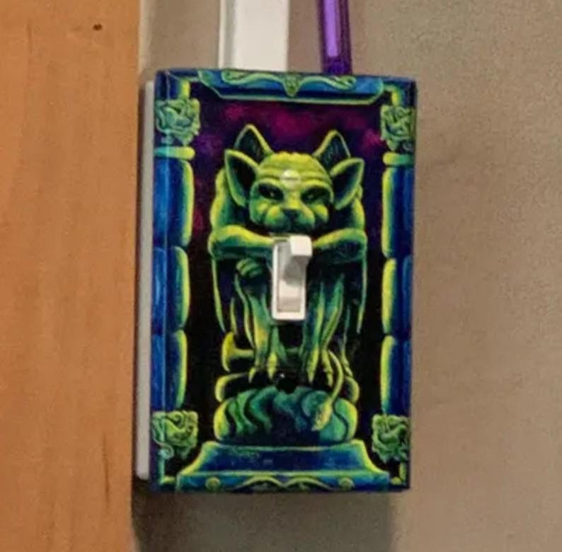 May include: A decorative light switch cover featuring a green gargoyle design against a purple and black background. The cover has a blue border with green accents and a white switch.