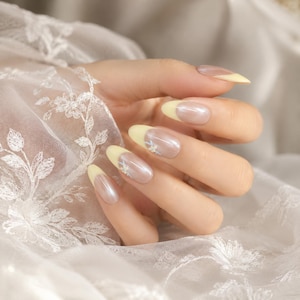 May include: Almond-shaped fingernails featuring a glossy, pearlescent nude base with pale yellow French tips. Each nail is accented with a small, delicate white and light blue floral design placed at the transition between the nude and yellow polish.