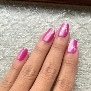 Può includere: Primo piano di una mano con quattro unghie dipinte di rosa acceso. Le unghie sono rettangolari e lucide, riflettono la luce. Lo sfondo è una superficie chiara e strutturata.