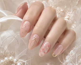 May include: A set of press-on nails featuring a soft pink base color. The design includes a gold quilted pattern with tiny studs, a delicate gold bow accent, small pink star motifs, and a gold metallic tip on the pinky nail for a refined look.