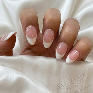 Op de afbeelding: Een hand met French manicure nagels. De nagels zijn ovaal van vorm met een lichtroze basis en witte punten. De tekst "AMBEREA ART STUDIO HANDCRAFTED LUXURY NAILS" is ook zichtbaar.