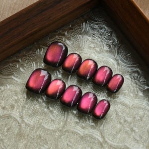 Op de afbeelding: Een set van tien persnagels met een glanzende afwerking. De nagels gaan over van een diepe bordeauxrode kleur naar een levendige oranjerode tint. De nagels zijn gerangschikt op een decoratief glazen oppervlak in een houten frame.