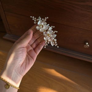 Könnte beinhalten: Ein goldfarbener Haarkamm mit zwei durchscheinenden weißen Blütenakzenten und goldfarbenen Staubgefäßen in der Mitte. Das Design ist mit kleinen weißen Kunstperlen und zarten goldfarbenen Drahtzweigen verziert, die ein florales Haaraccessoire bilden.
