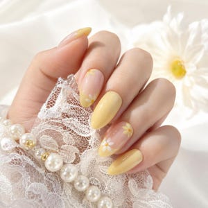 Può includere: Unghie press-on a forma di mandorla con un colore giallo tenue e accenti trasparenti decorati con piccoli motivi a margherita bianca. Il set include un delicato polsino in pizzo bianco ornato da una fila di perle bianche e perline dorate.