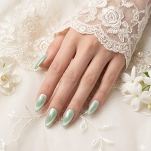 May include: A hand with long, oval-shaped, pale green nails. The nails have a pearlescent finish. The hand rests on white, lacy fabric with floral embroidery. A white lace sleeve and white flowers are also visible.