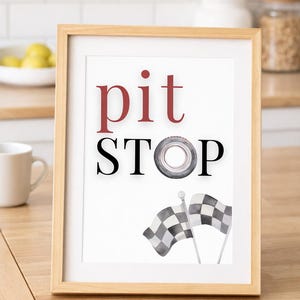 May include: A framed print with the words "pit STOP" in red and black, with a tire graphic replacing the "O". Two watercolor checkered flags are at the bottom. The frame is light wood.