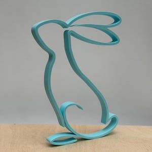 Könnte beinhalten: Hellblauer, 3D-gedruckter Hasen-Ausstecher. Der Ausstecher ist eine durchgehende Linie, wobei der Hase aufrecht sitzt. Der Ausstecher ist aus Kunststoff und steht auf einer Holzoberfläche.
