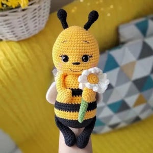 May include: A handmade crochet bee doll in yellow and black stripes, holding a white daisy. The bee has black antennae, wings, and legs. The doll is approximately 15 cm tall.
