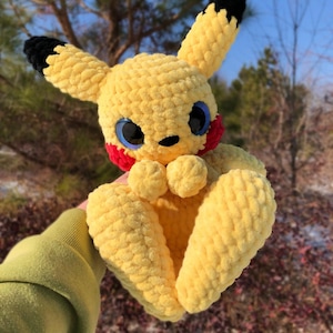 Może przedstawiać: Ręcznie robiona żółta pluszowa zabawka Pikachu z czarnymi końcówkami uszu, czerwonymi policzkami i dużymi niebieskimi oczami. Plusz wykonany jest z miękkiego, teksturowanego materiału i ma uśmiechniętą minę. Zabawka jest popularną postacią z serii Pokémon.
