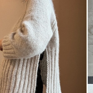 May include: A light grey, oversized knitted cardigan with long sleeves. The cardigan features a ribbed texture on the front and a softer, more textured knit on the sleeves. The garment is open at the front and appears to be made of a soft, warm material.