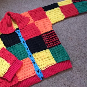 Op de afbeelding: Een kleurrijk, handgehaakt vest met een patchwork-ontwerp. De trui heeft een verscheidenheid aan heldere kleuren, waaronder rode, gele, oranje, groene en zwarte vierkanten. Het heeft een rode kraag, blauwe knoopbies en zwarte knopen.