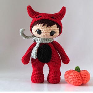 May include: A handmade crochet doll dressed in a red devil costume with horns, a black belly, and a gray scarf. A small orange crochet pumpkin with a green stem sits beside the doll. The doll has brown hair and black eyes.