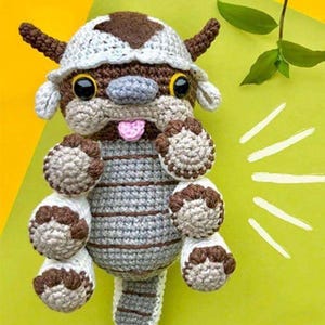 May include: Crocheted plush toy in the shape of a cartoon character. The toy features a brown and white head with large black eyes, small ears, and a pink tongue. The body is gray with brown stripes, and the paws are white and brown.